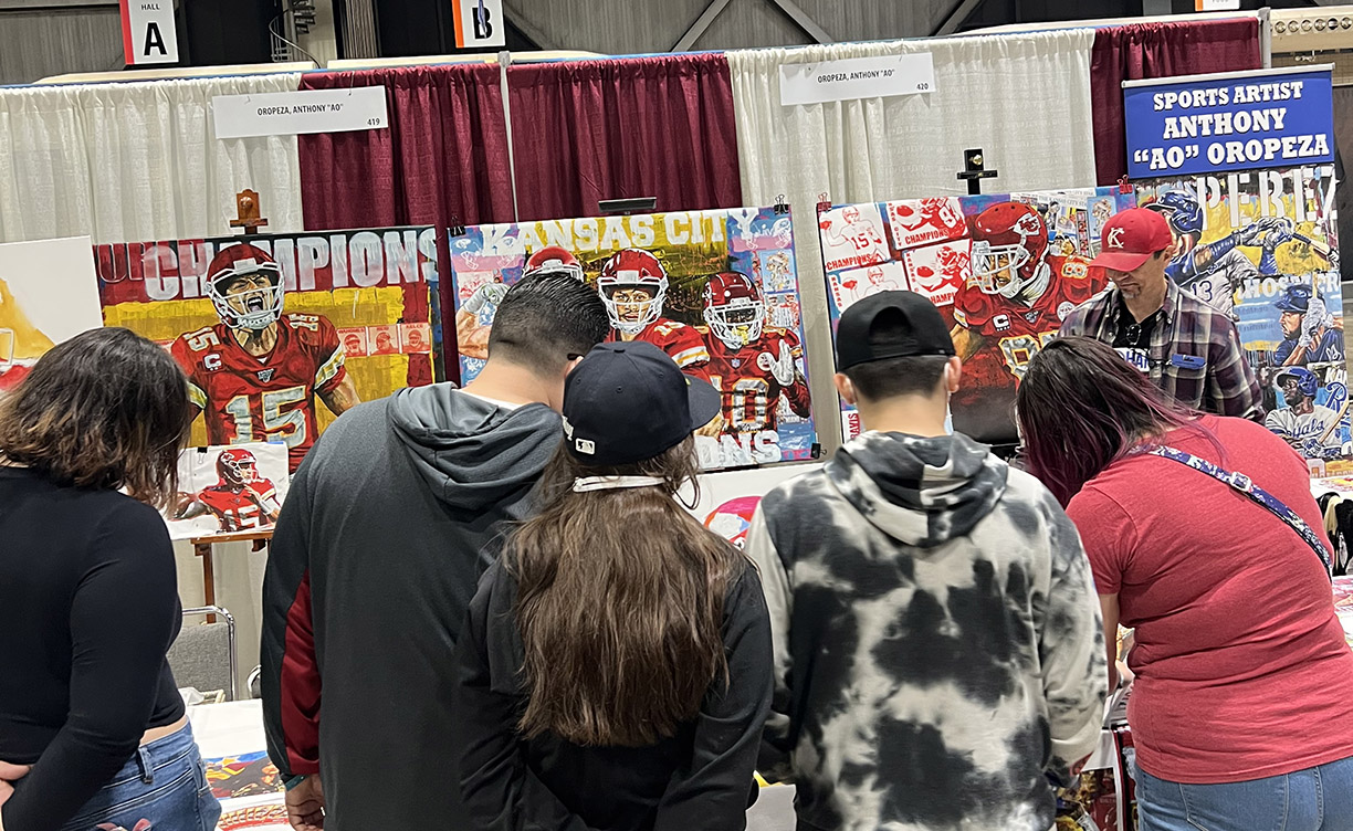 Sports artist Anthony Ao Oropeza at his booth at the KC Comicon with several onlookers checking out his baseball and football sports paintings.