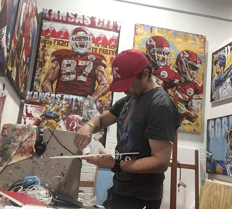 Sports artist Anthony AO Oropeza painting in his studio, surrounded by his original Chiefs and Royals paintings
